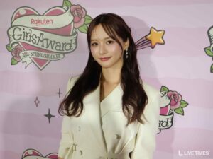 Young woman posing in front of a pink illustrated backdrop at a fashion event, wearing a cream blazer and pearl earrings.