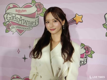 Young woman posing in front of a pink illustrated backdrop at a fashion event, wearing a cream blazer and pearl earrings.