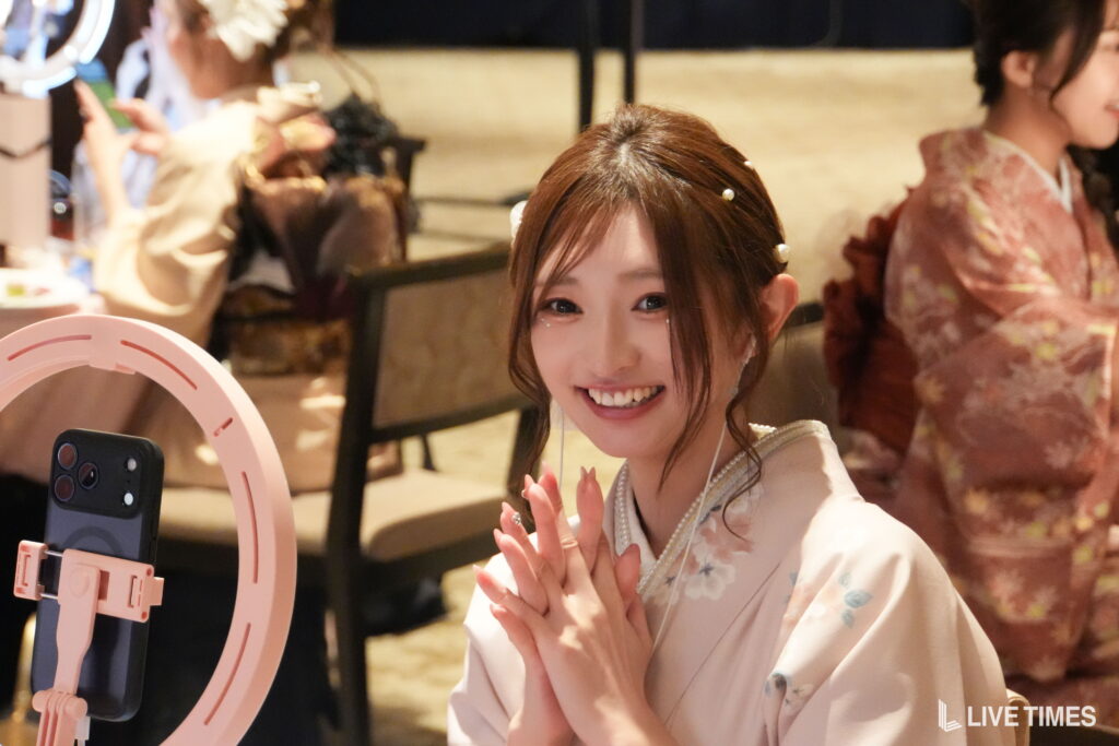 Smiling young woman in a light kimono with floral pattern posing for a selfie, hands raised near her face, pink ring light and phone visible in the foreground.