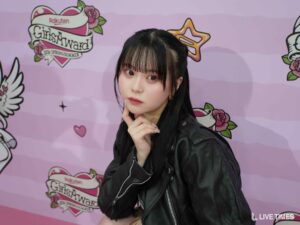 Young woman posing against a pink backdrop with 'GirlsAward' branding, wearing a black jacket and touching her chin thoughtfully.