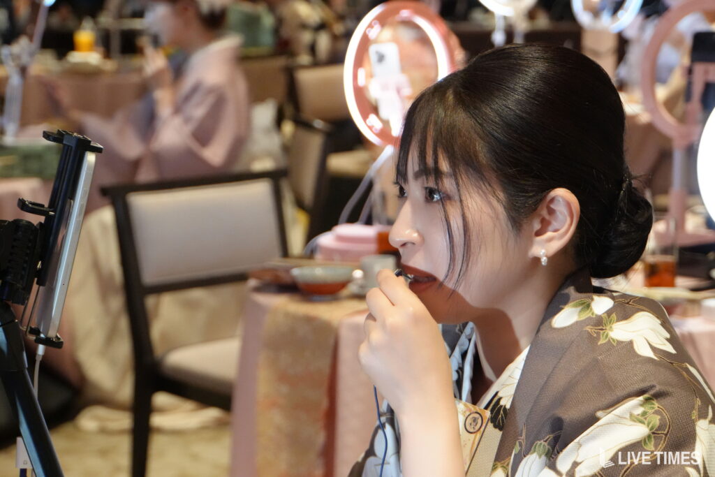 Profile view of a woman wearing a floral kimono applying lipstick while looking at a screen on a stand, with a ring light in the background.