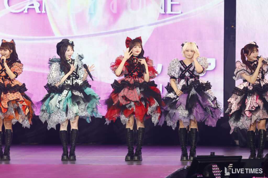 Five performers in colorful, layered dresses with boots sing on a pink-lit stage, each holding a microphone.