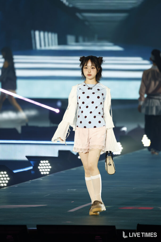 Young performer on a lit stage wearing a light blue polka-dot top and pale pink shorts, carrying a beige bag.