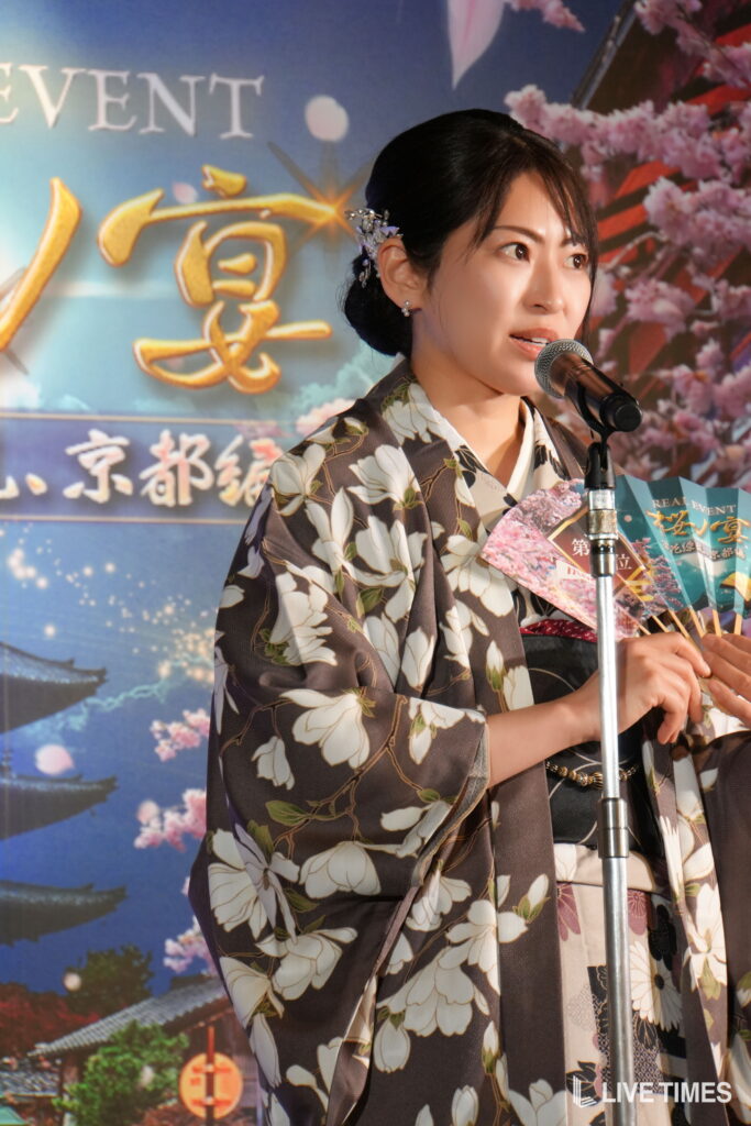 Woman in a floral kimono speaks into a microphone on stage, with a cherry blossom backdrop and event banners behind her.