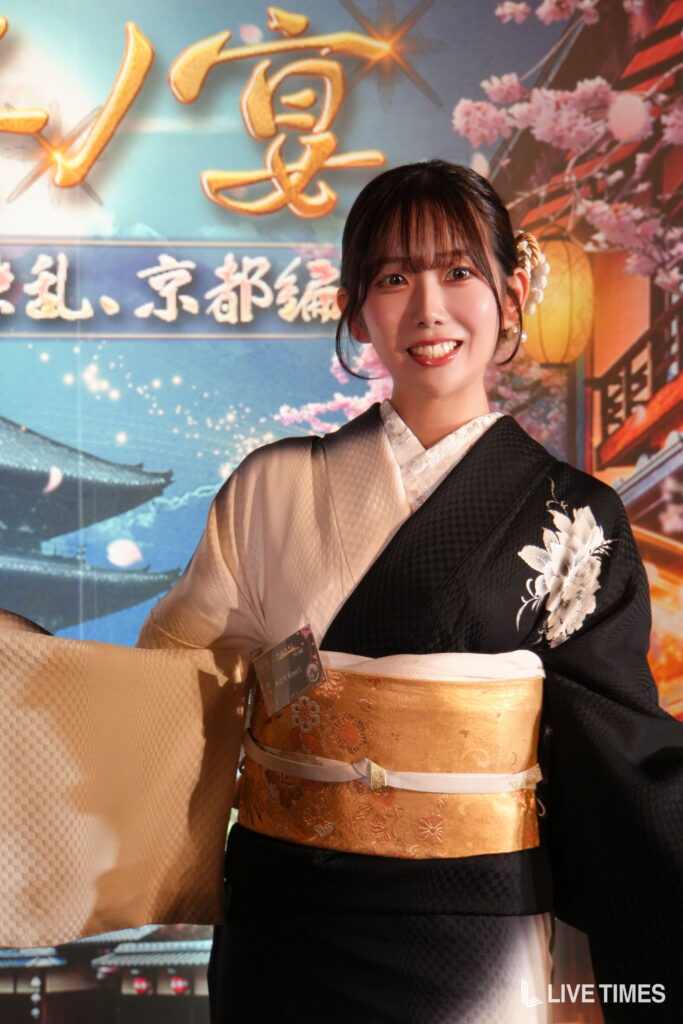 Young woman in a black and cream kimono smiling at camera at a Japanese festival backdrop with cherry blossoms and lanterns