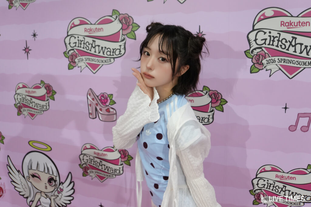 Young woman posing with her hand near her chin in front of a pink backdrop with GirlsAward logos and decorations (Rakuten).