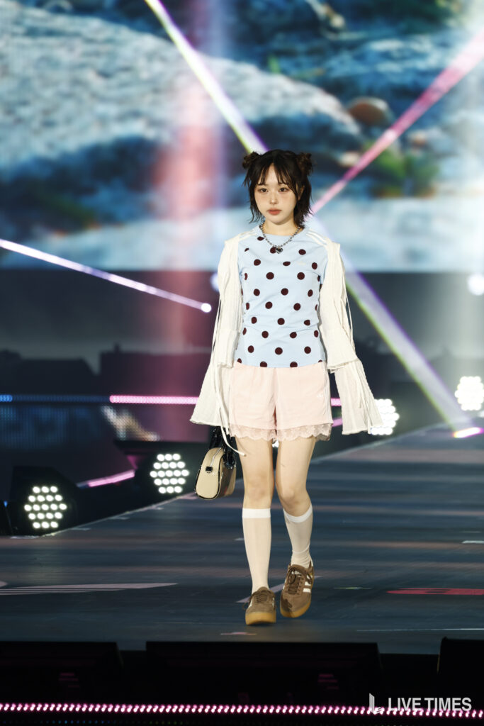 Young model walking a runway in a light blue polka-dot top, pale pink shorts, white sheer sleeves, and a small beige handbag.