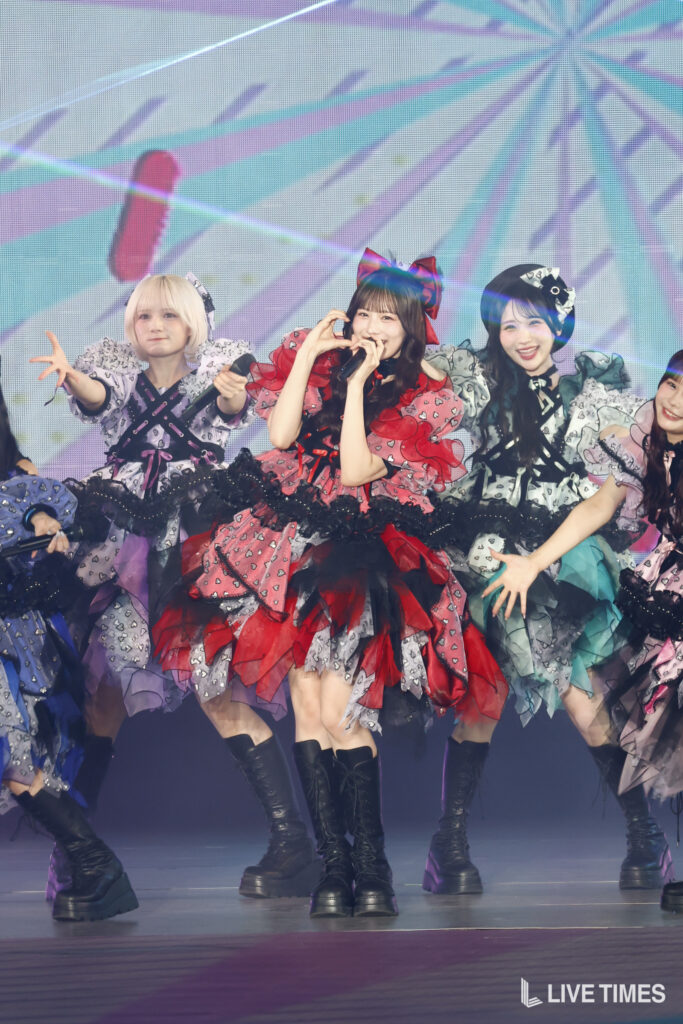 Group of female performers in colorful frilly costumes on stage; center singer makes a heart gesture with her hands while singing.
