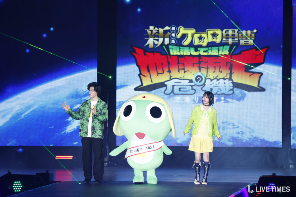 Two hosts stand on a lit stage beside a large green cartoon mascot with oversized eyes, against a space-themed backdrop.