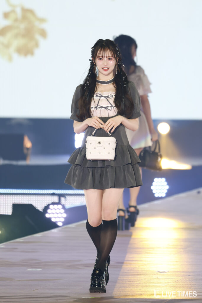 Runway model in a gray layered dress, carrying a white studded handbag and wearing black knee-high socks and glossy shoes.