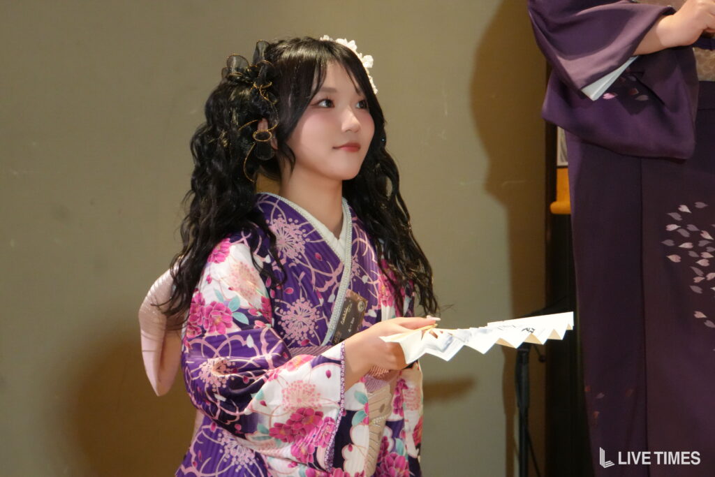 Young woman in a purple floral kimono on stage, looking to the side while holding papers.
