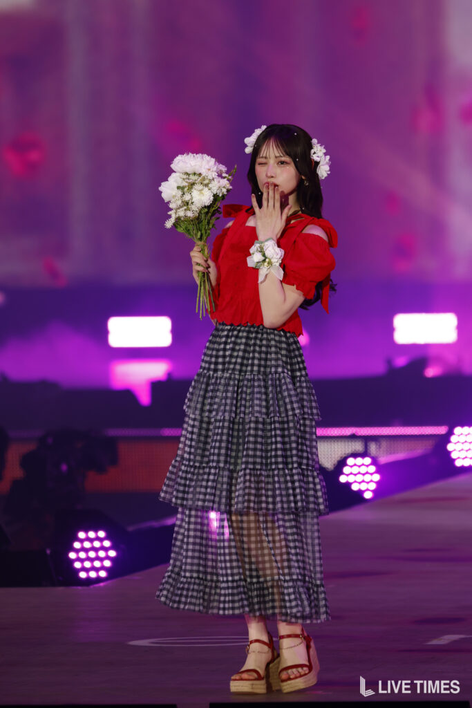 Young woman in a red blouse and tiered plaid skirt holds a bouquet of white flowers on stage, wearing a flower crown.