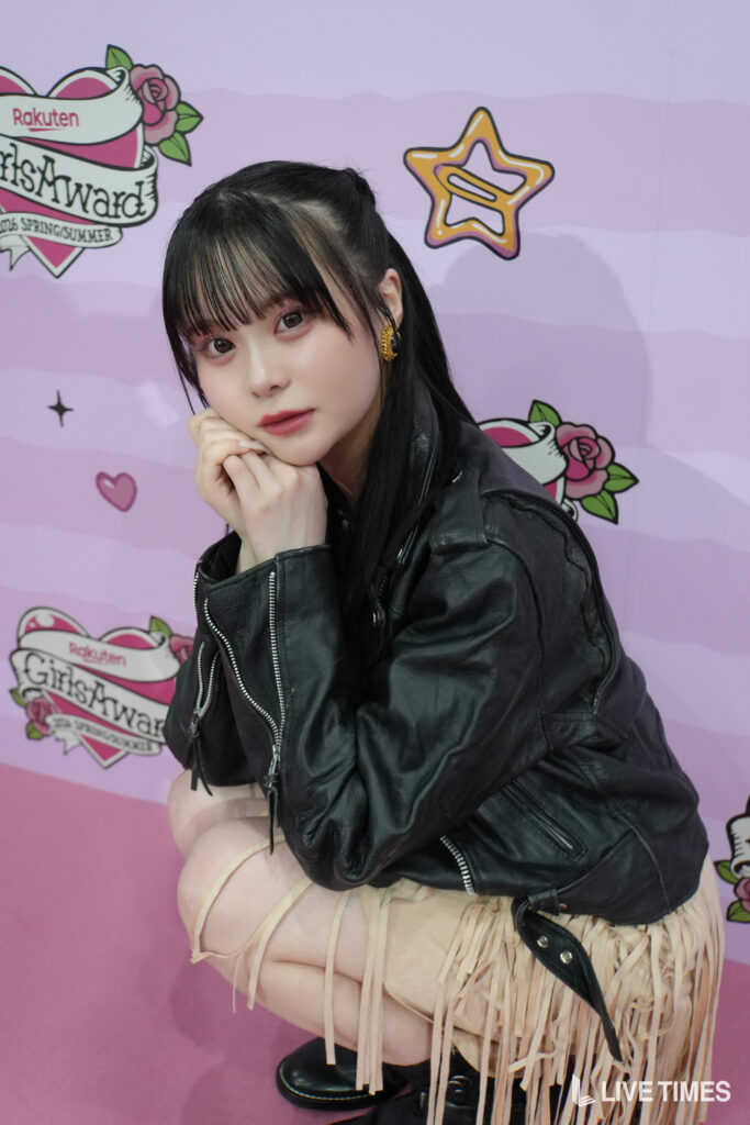 Young woman with black hair and bangs resting her chin on her hands, posing against a pink backdrop with fashion logos behind her.