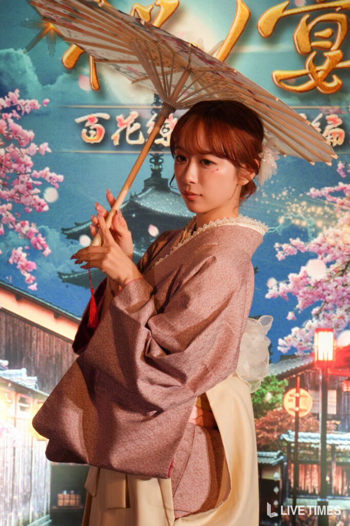 Woman in a pink kimono holding a traditional paper parasol, posed before a colorful Japanese backdrop with cherry blossoms and a temple.