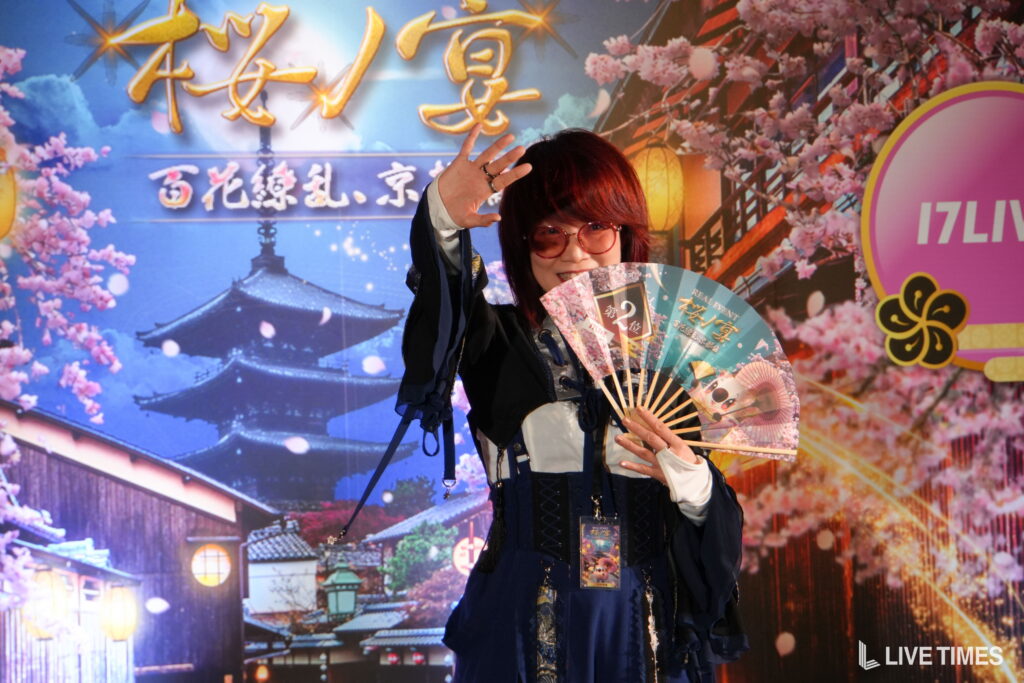 Person wearing dark clothing and red glasses waves with a decorative fan at a colorful festival backdrop with cherry blossoms and lanterns.