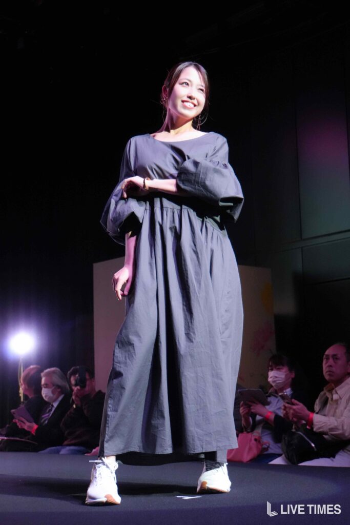Woman on stage smiling, wearing a loose gray dress and white sneakers, performing with audience in the background.