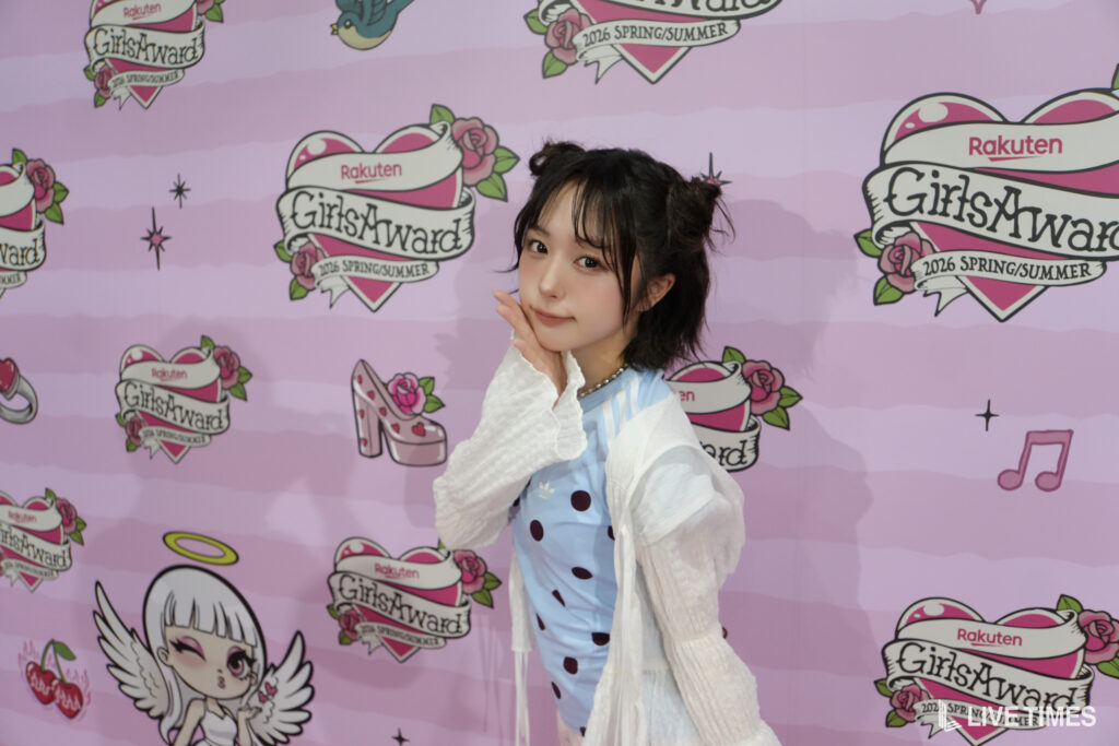 Young woman posing with her hand near her chin in a light blue polka‑dot dress and white cardigan, in front of a pink backdrop with 'GirlsAward' logos and cartoons.