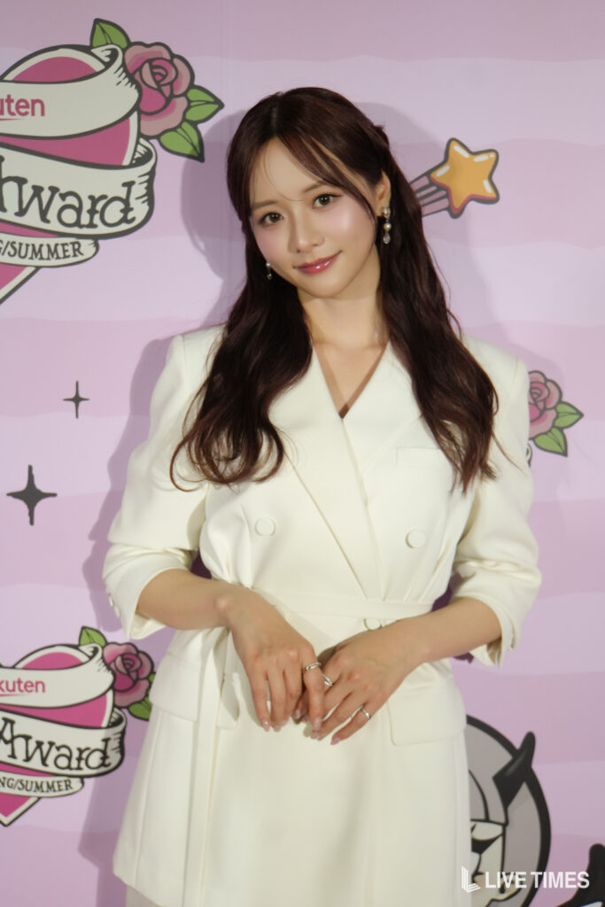 Portrait of a woman with long dark hair wearing a white blazer, smiling at the camera against a pink event backdrop with logos.