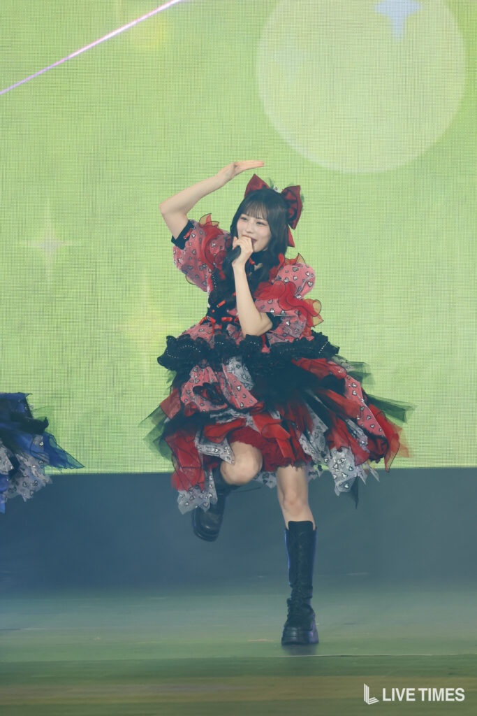 Performer in a red and black frilly dress with a large bow, singing into a microphone on stage with a green backdrop.