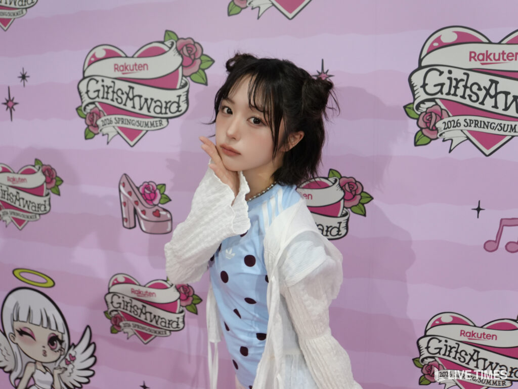 Young woman posing in front of a pink GirlsAward backdrop with logos and roses, wearing a light blue polka-dot dress and white cardigan.