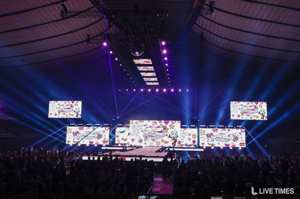 Concert stage in a large, dark arena with purple and blue spotlights and multiple LED screens displaying colorful graphics and 'GirlsAward' branding.