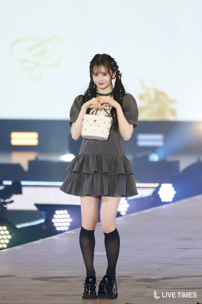 Young woman on a stage posing with a white studded handbag, wearing a gray tiered dress and black knee-high socks.