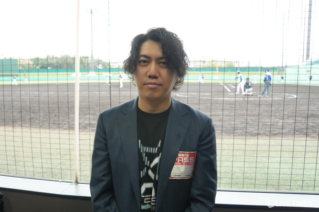 Man in a dark blazer standing indoors in front of a chain-link fence at a baseball field, with players visible in the background.