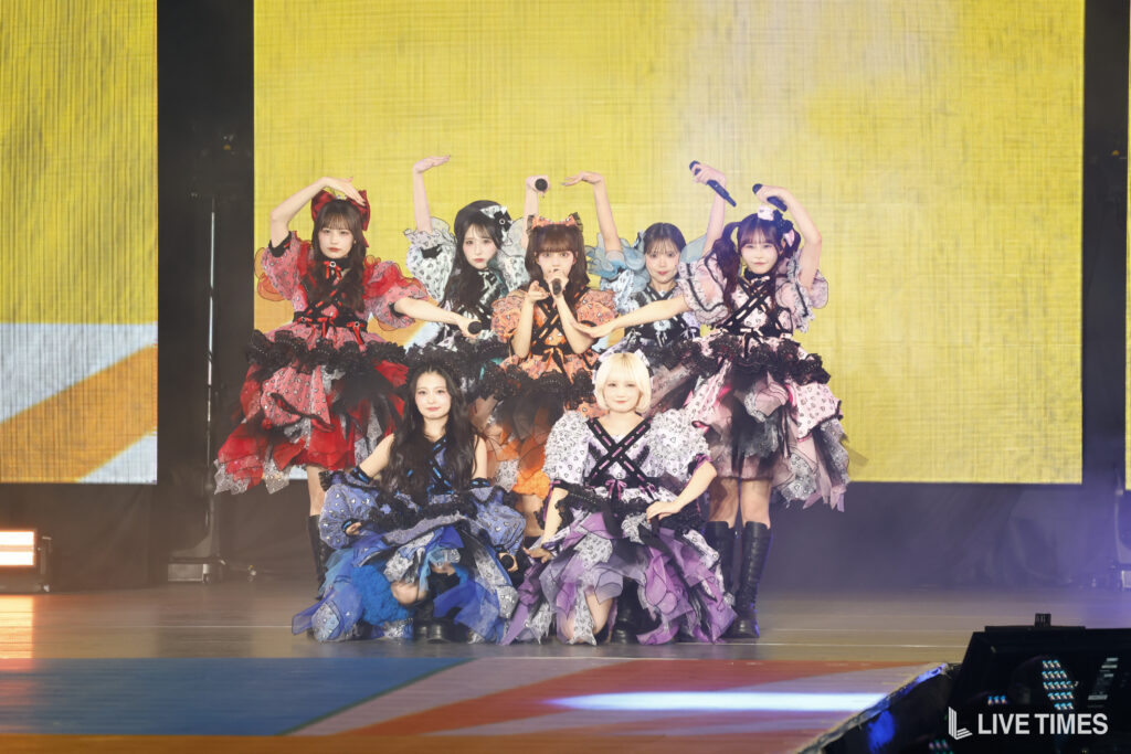 Group of female performers in colorful frilly outfits posing on stage during a show, with yellow backdrop and lights in the background.