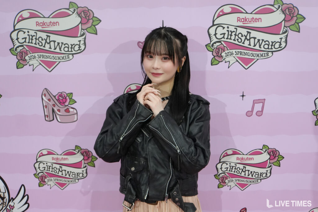 Young woman in a black leather jacket posing with hands clasped in front of a pink GirlsAward backdrop.