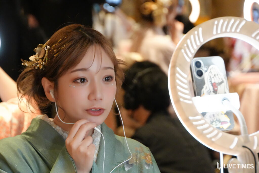Young woman in a green traditional outfit with gold hair ornaments, wearing white earbuds, looking to the side near a circular vanity light.