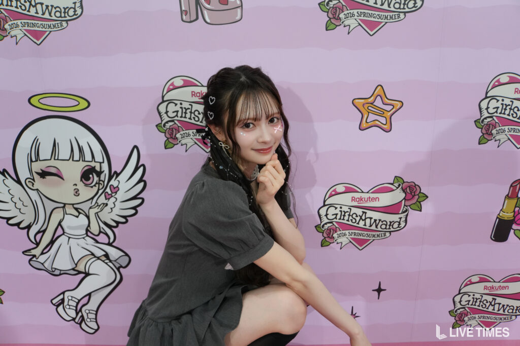 Young woman posing with hands under her chin in front of a pink event backdrop featuring cartoon angels and 'GirlsWard' logos.