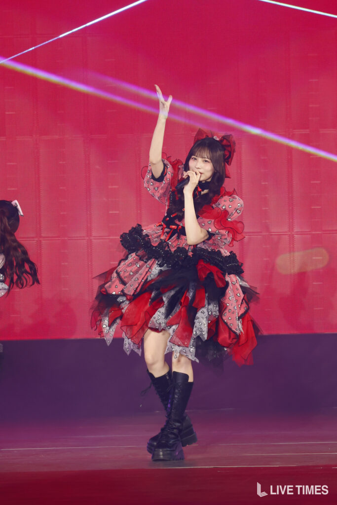 Performer in a red-and-black frilly stage costume singing into a microphone with one arm raised on a pink-lit stage.
