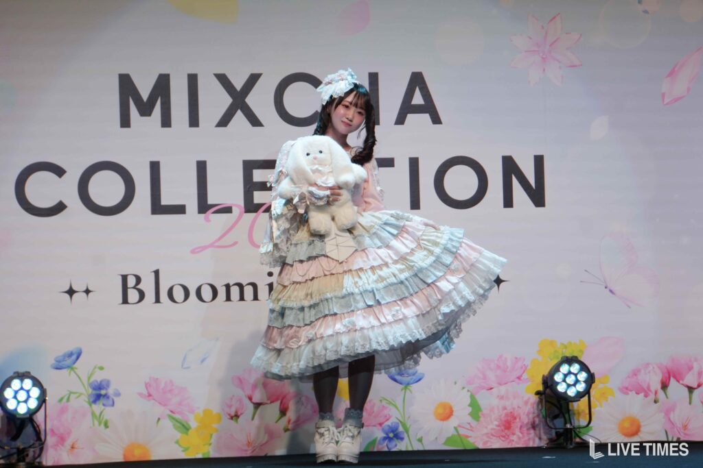 Cosplay model in a pastel frilly dress posing with a white plush bunny on stage with a floral backdrop and 'MIXCOLLECTION' text.