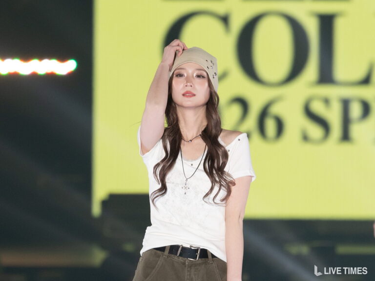 Woman on stage adjusting a beige beanie in front of a bright yellow backdrop with a LIVE TIMES logo at the bottom right.