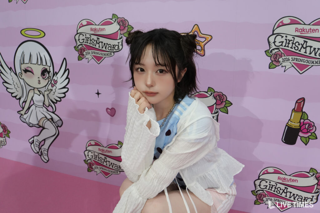 Young woman posing with her chin resting on her hand in front of a pink GirlsAward backdrop filled with logos and illustrations of angels and roses.