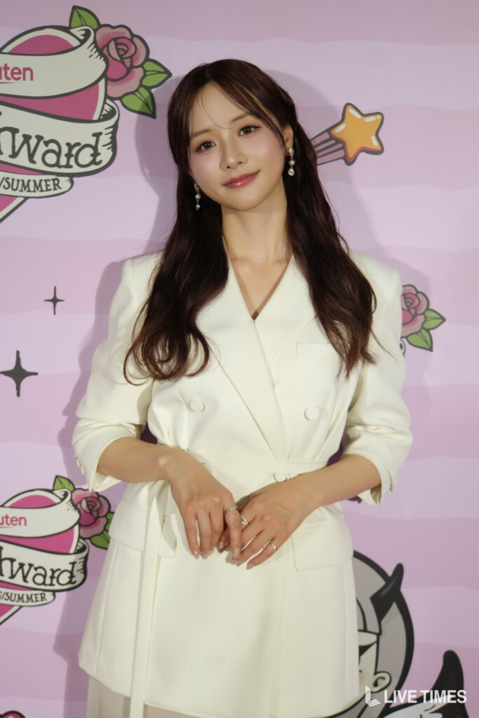 Woman in a white blazer posing at a pink event backdrop with cartoon graphics and a watermark at the bottom corner.