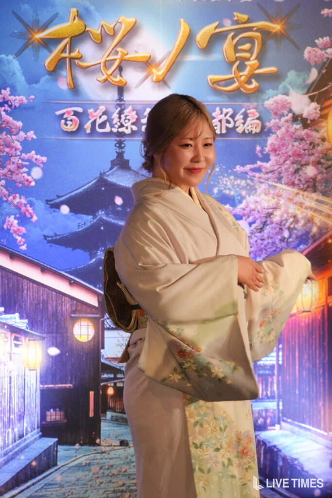 Woman in a pale kimono smiling in front of a colorful cherry blossom backdrop with Japanese script and lanterns on the set.