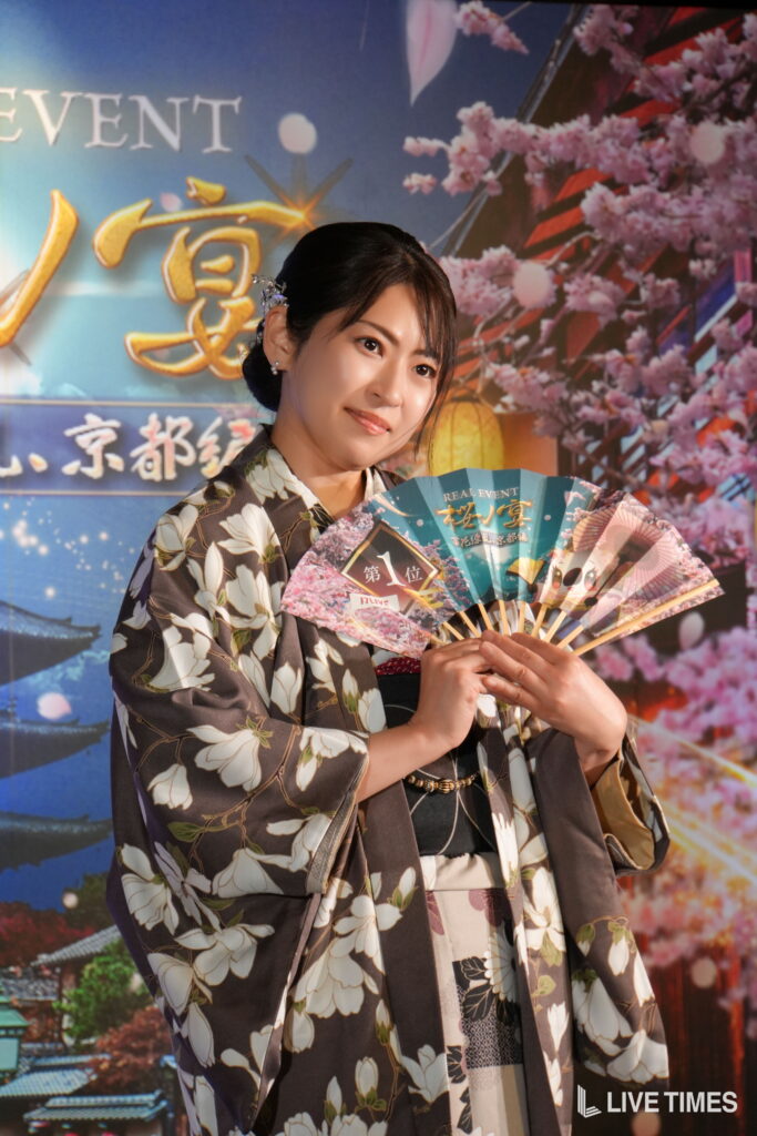 Smiling woman in a floral kimono holding a decorative folding fan at a festival with cherry blossom backdrop behind her.
