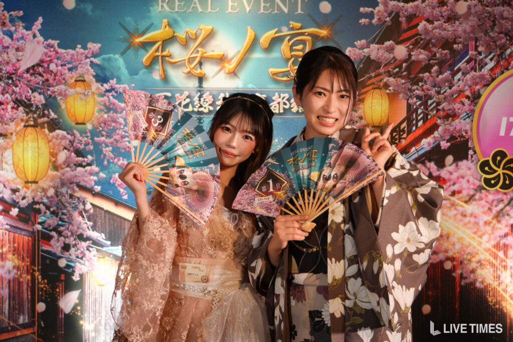 Two women in floral kimonos posing with decorative fans in front of a cherry blossoms and festival backdrop