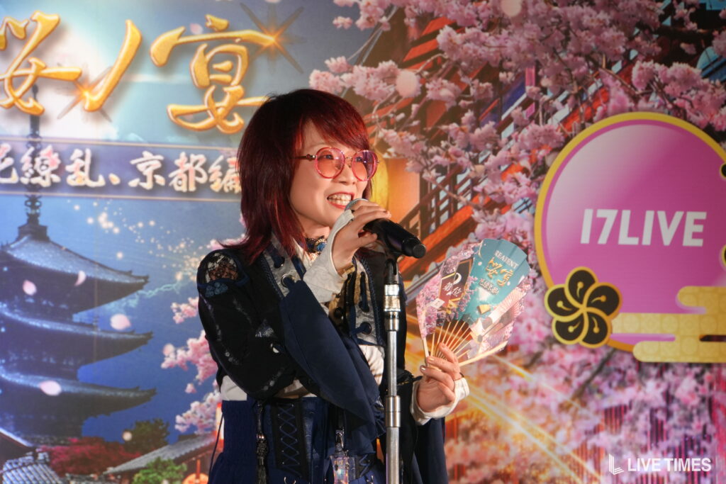 Female singer with red hair and pink glasses performing into a microphone on a colorful stage backdrop filled with cherry blossoms and signage.