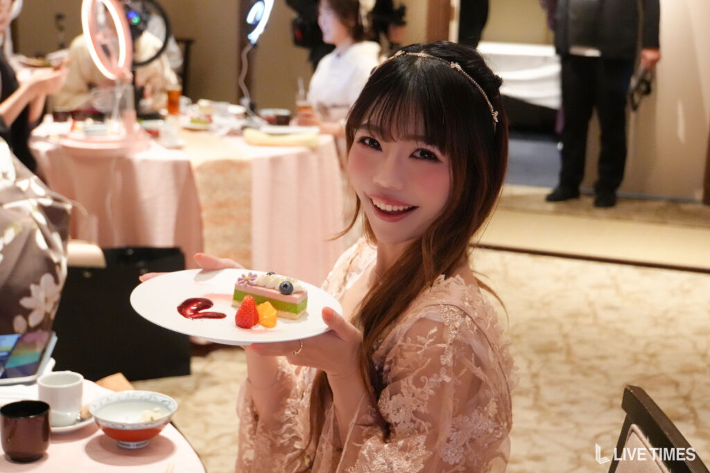 Smiling woman in pastel lace dress presenting a plate with small layered cake and fruit at a party tablescape.