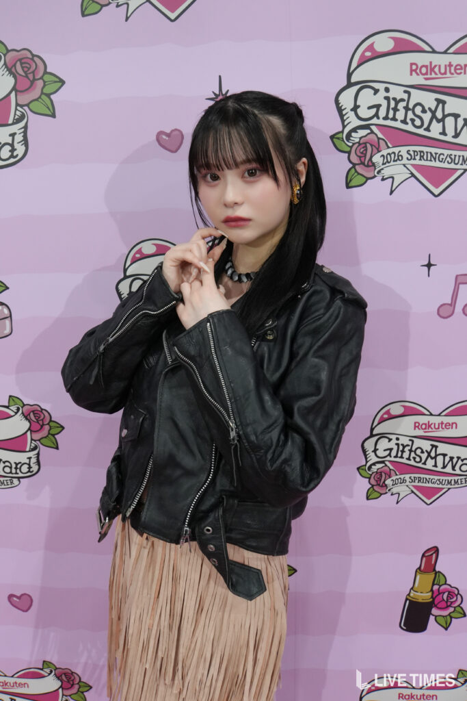Young woman with black hair wearing a black leather jacket and beige fringe skirt, posing with hands near her chin in front of a pink GirlsAward backdrop.
