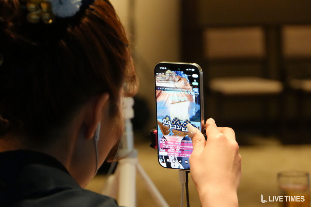 Close-up of a hand holding a smartphone showing a colorful game interface, with a ship-themed scene and a Live Times watermark bottom-right.