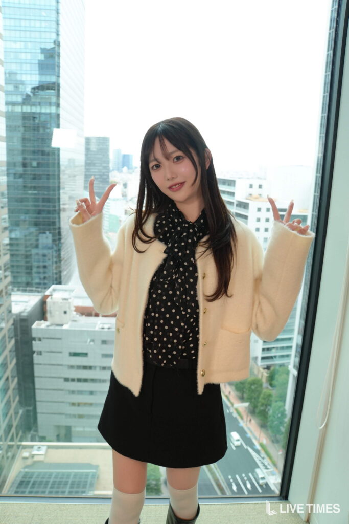 Young woman posing with peace signs by a large window in a high-rise, cityscape behind her.
