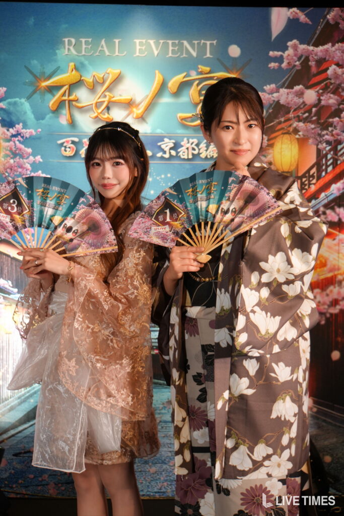 Two women in ornate kimonis pose with decorative fans at a festival backdrop.