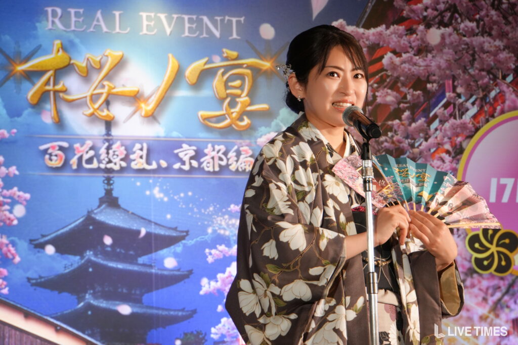 Woman in a floral kimono speaking into a microphone on a stage with a blue backdrop and cherry blossoms behind her.