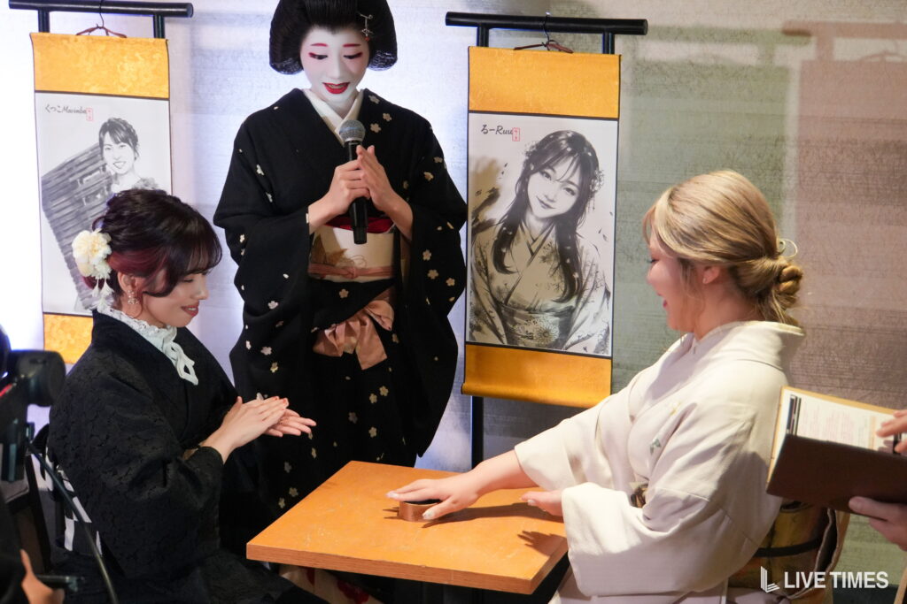 Geisha in a black kimono holds a microphone while two seated women in kimonos clap at a wooden table, with orange banners and portraits behind.