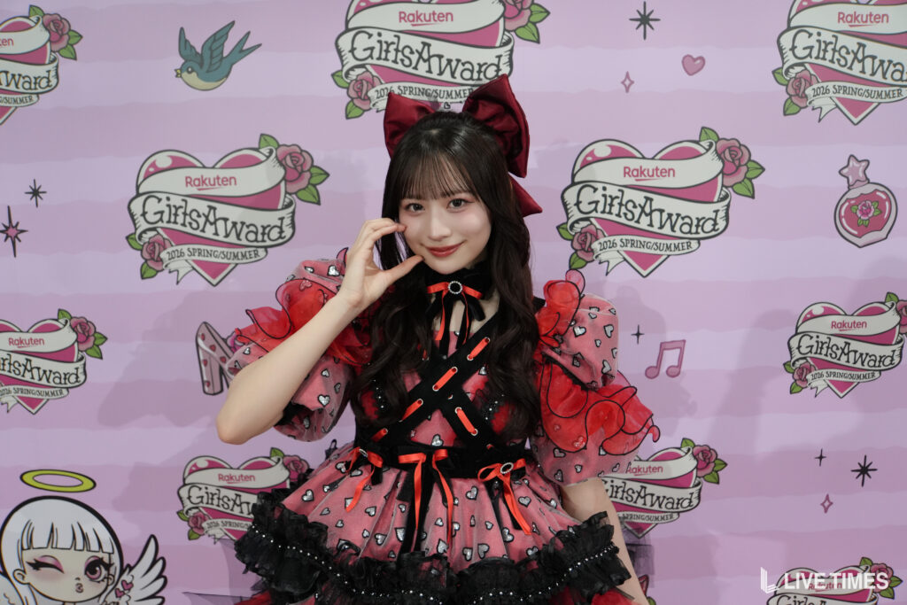 Young woman in a red and black cosplay dress with a large red bow posing and smiling in front of a pink GirlsAward backdrop.