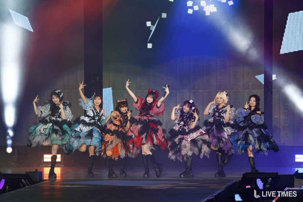 Eight performers in colorful, ruffled dresses dance in unison on a lit stage with blue and red spotlights and a digital backdrop behind them.