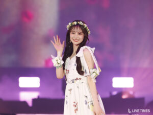 Woman in a white floral dress waving from a purple-lit stage, wearing a flower crown and wrist corsage.